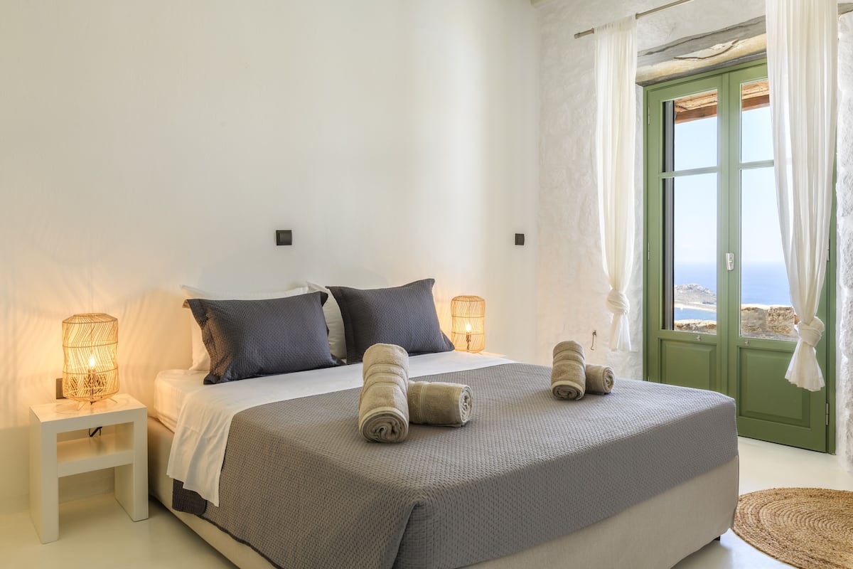 Une chambre à coucher avec un lit, des oreillers, une table de chevet avec une lampe, et une vue sur l’extérieur, visible à travers une porte verte.