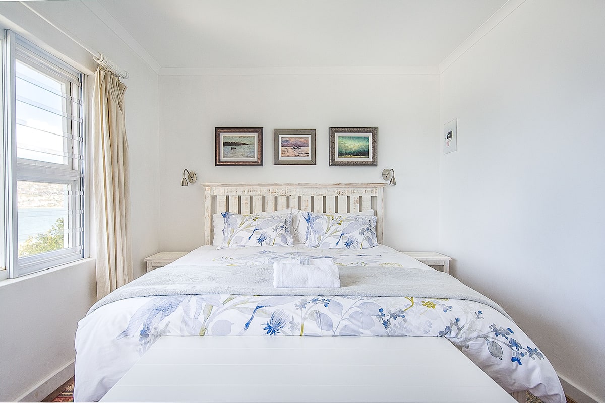 A bedroom with a bed, a nightstand, a window with curtains, and three framed pictures on the wall.