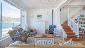 Een ruime woonkamer met een groot raam met uitzicht op een strand, een houten trap en een boekenkast.