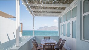 Een overdekt terras met een tafel en stoelen, met uitzicht op zee en bergen.