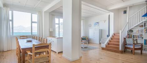 A spacious living area with a wooden dining table, chairs, a staircase, and a bookshelf.