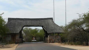 Una entrada con techo de paja, paredes de piedra, banderas y un camino que se adentra en un paisaje natural.