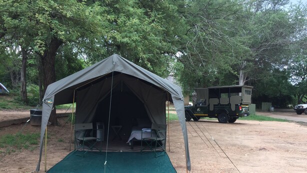 A tent with two beds and a table, set up on a dirt ground with trees in the background. - Kruger Park Tours (Mbombela)