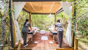 A wooden deck with two massage tables, a white table, and a hanging light fixture. - Kruger Park Tours (Mbombela)