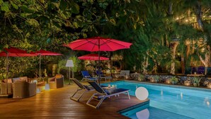 Um deck de madeira com espreguiçadeiras e guarda-sóis vermelhos, uma piscina e vegetação ao redor.