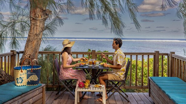 Um deck de madeira com mesa e cadeiras, com vista para uma praia, oceano e palmeiras.