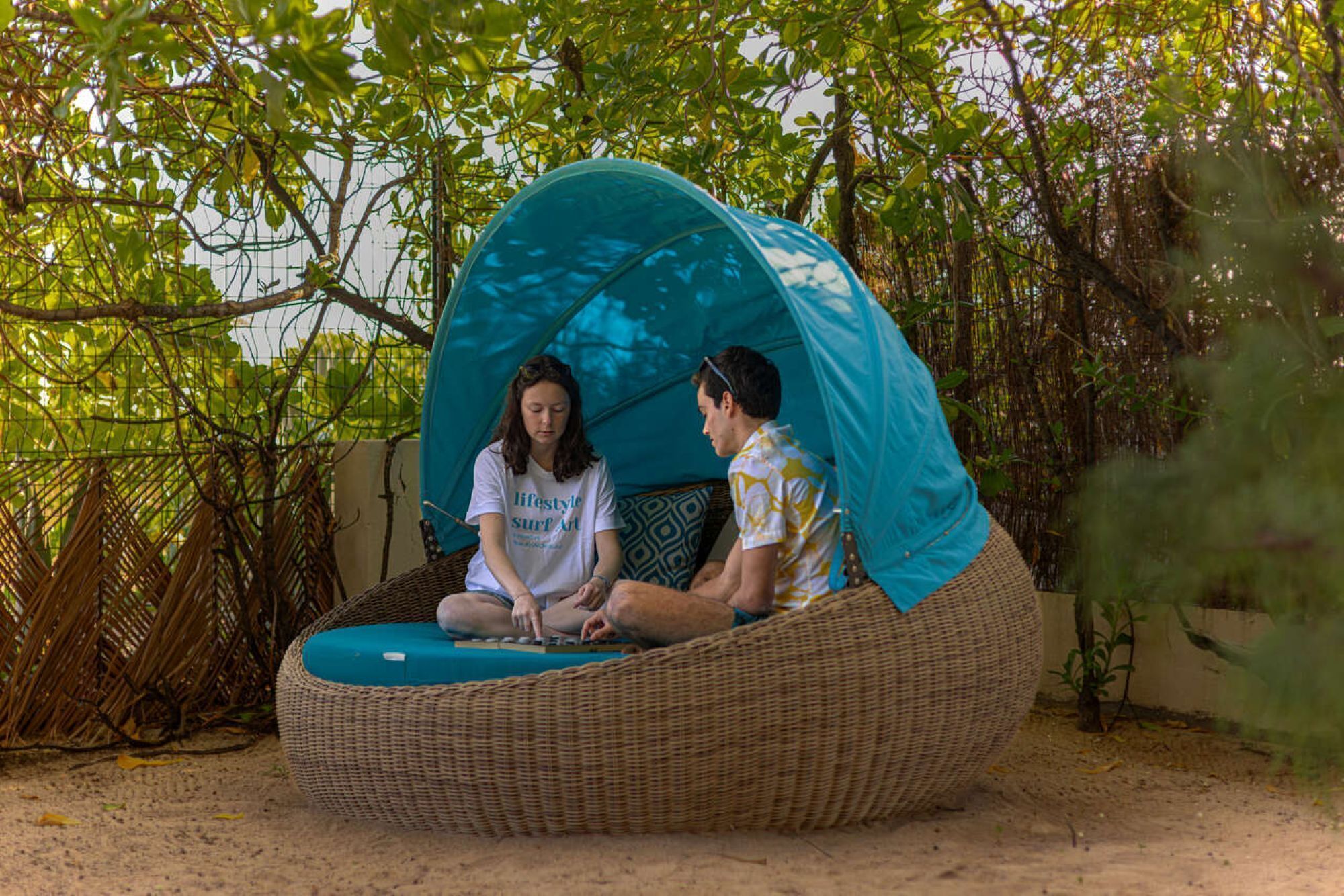 Two people sitting in a woven, round outdoor seating area with a blue canopy.