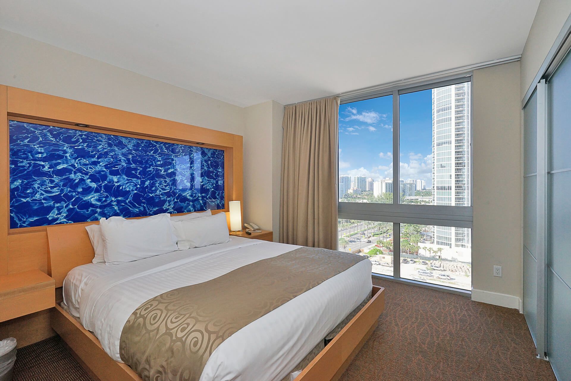 Quarto de hotel com uma cama grande, vista da cidade através de janelas amplas e uma obra de arte azul na parede.