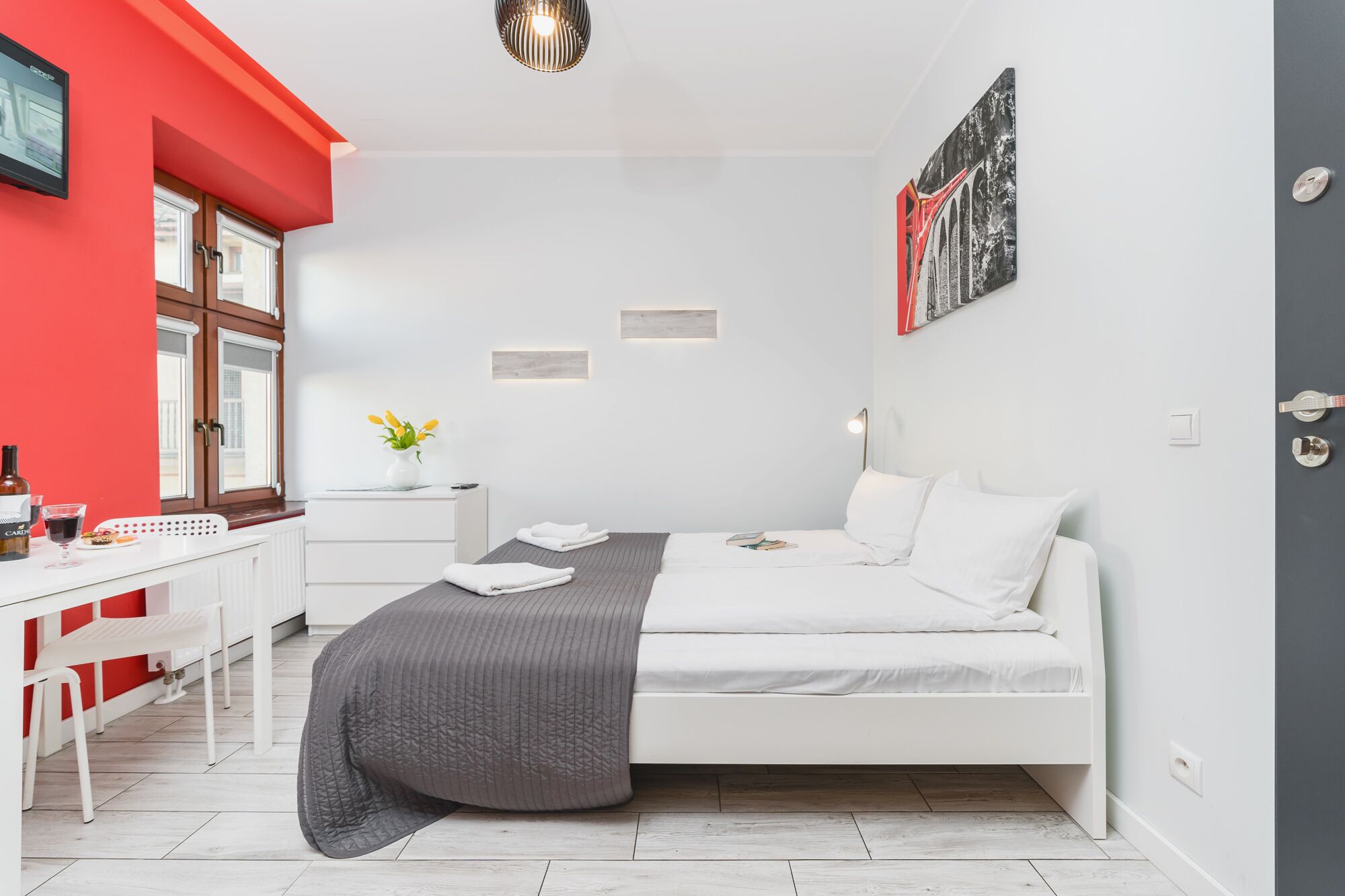 A modern bedroom with a white bed, a desk with a chair, a red accent wall, and a painting on the wall.