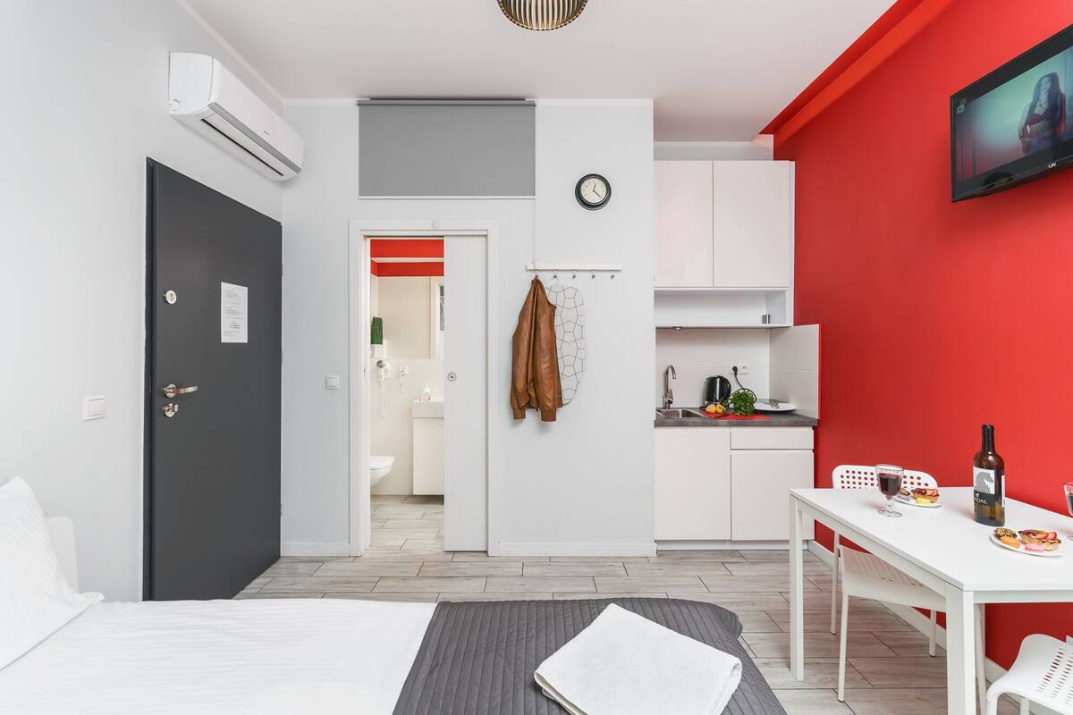 A modern hotel room with a bed, a desk, a dining table, a kitchen area, and a TV mounted on the wall.