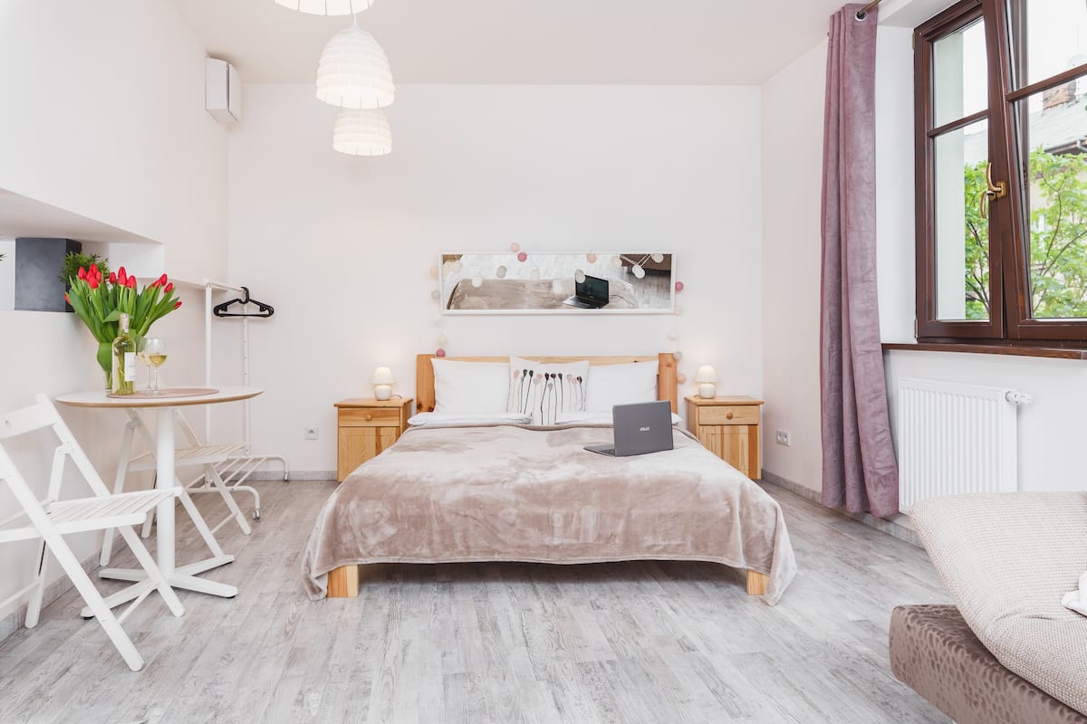Une chambre à coucher avec un lit, un bureau et une chaise. Il y a une fenêtre avec des rideaux, un radiateur et une œuvre d’art accrochée au mur.