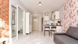 Una sala moderna con un sofá gris, una mesa de centro blanca y una zona de cocina con armarios blancos y una escalera de madera.