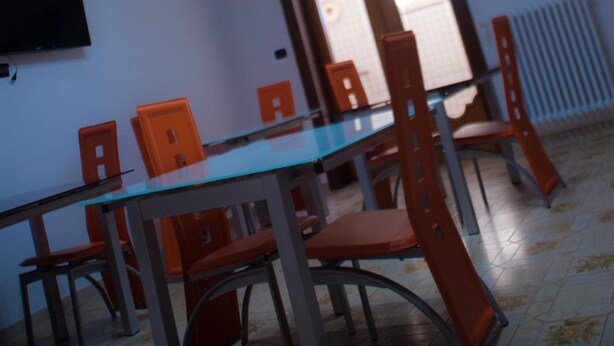 Zona pranzo con tavolo in vetro e sedie arancioni, televisore a parete e porta che conduce in un'altra stanza.