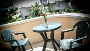 Un balcone con due sedie, un tavolo, un vaso con un fiore e vista su edifici e palme.