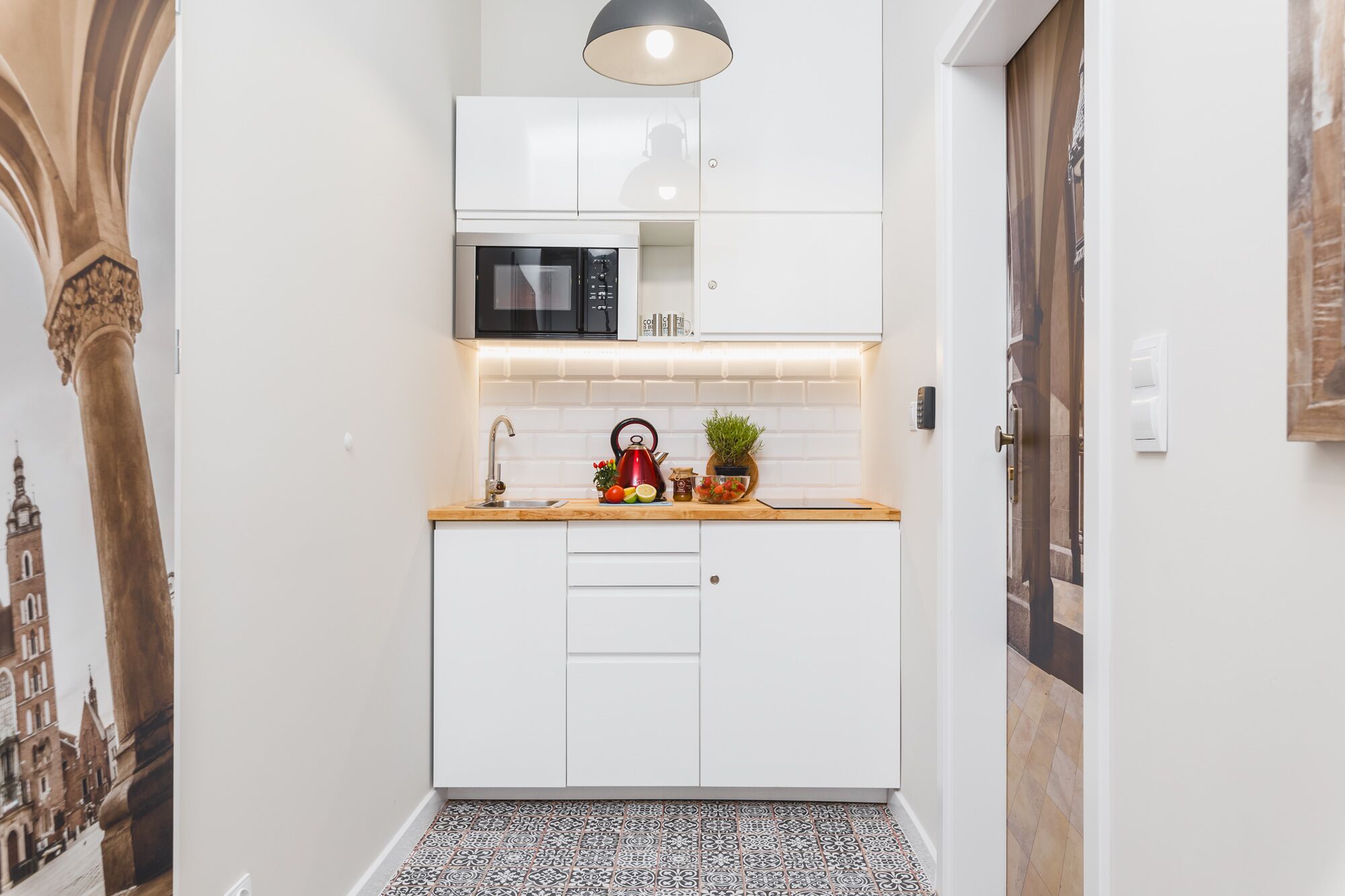 Cocina compacta con armarios blancos, un microondas, un lavamanos y una pequeña encimera con una tetera y productos frescos.