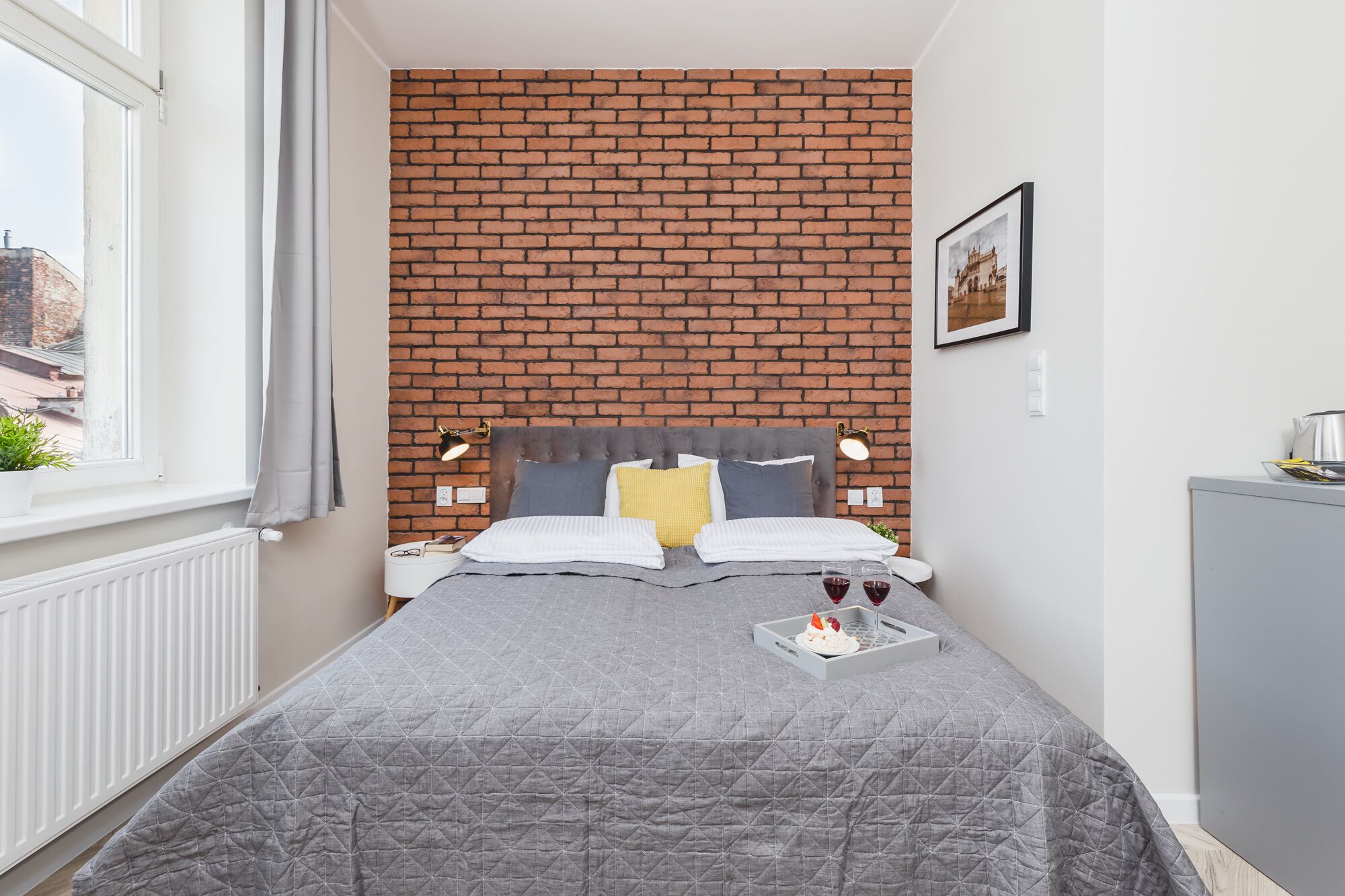 Un dormitorio con pared de ladrillo, una cama con colcha gris, dos lámparas de noche, una mesita con una bandeja de copas de vino y un pastel, y un cuadro en la pared.