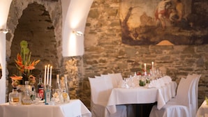 A restaurant with stone walls and a mural, tables set with white tablecloths and candles, and a vase with flowers on the left. - Residenz Schloss Engers (Neuwied)