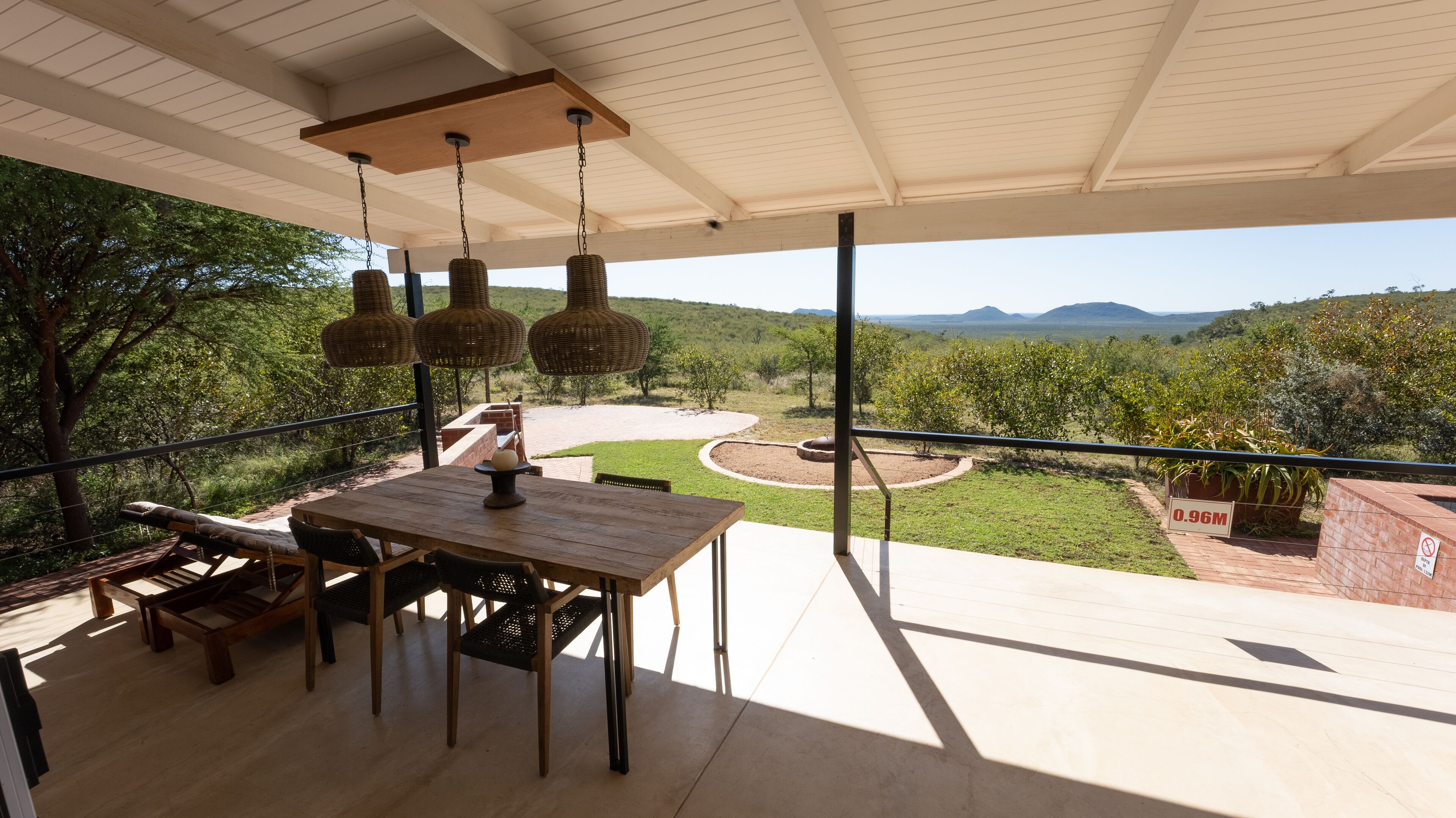 Un balcón con mesa y sillas de madera, una lámpara colgante y vistas a un jardín ajardinado y colinas distantes.