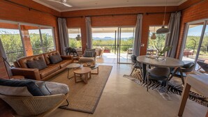 Amplia sala de estar con un sofá de cuero, sillas de mimbre, una mesa redonda y vistas al exterior a través de grandes puertas correderas.