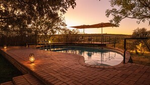 Un área de piscina con diseño circular, rodeada por un deck de ladrillo y un deck de madera con sillas y una sombrilla.