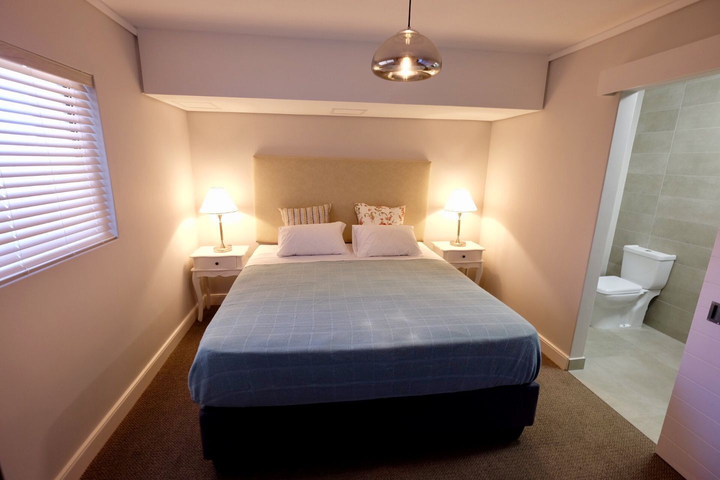A bedroom with a bed, two bedside tables with lamps, a window with blinds, and a bathroom with a toilet and shower.