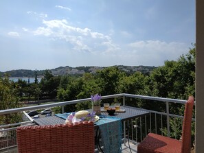 A balcony with a table set for two, overlooking a scenic view of a coastal town and hills. - Lilly Apartments (Vari-Voula-Vouliagmeni)