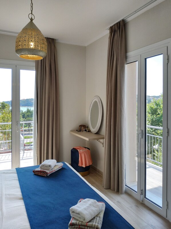 A bedroom with a bed, a bedside table, a mirror, and a view of the outdoors. - Lilly Apartments (Vari-Voula-Vouliagmeni)