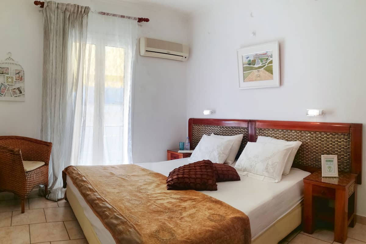 A hotel room with a large bed, a wooden headboard, a wicker chair, a nightstand, and a wall-mounted air conditioner.