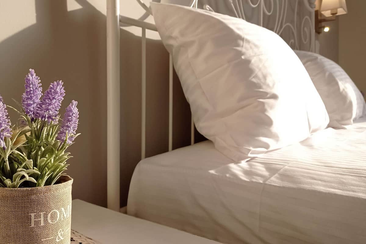 A neatly made bed with white linens and pillows, a woven basket with lavender plants, and a wall-mounted light fixture.