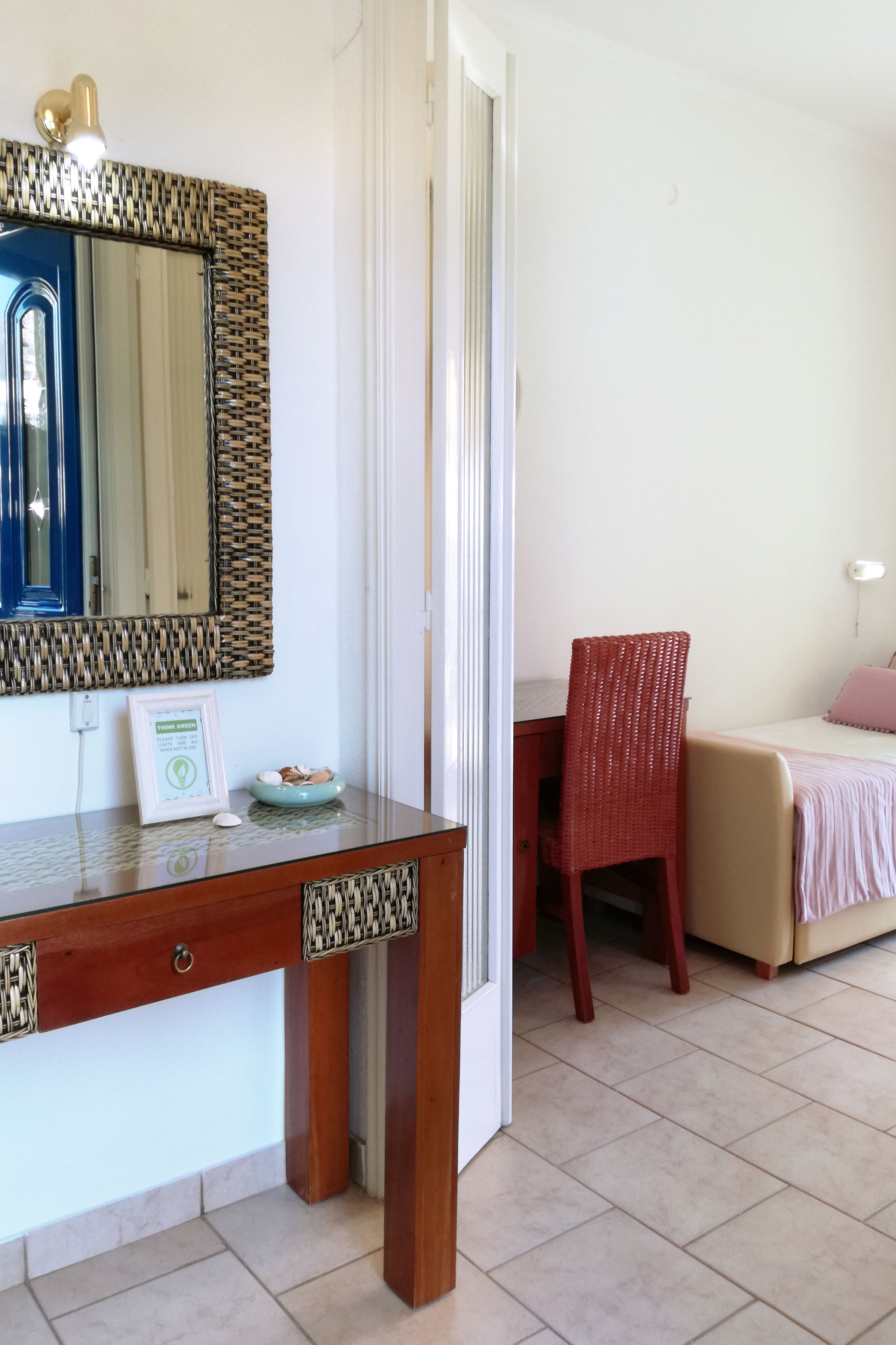 A hotel room with a wooden desk, a red chair, a bed with a pink pillow, and a mirror with a woven frame.