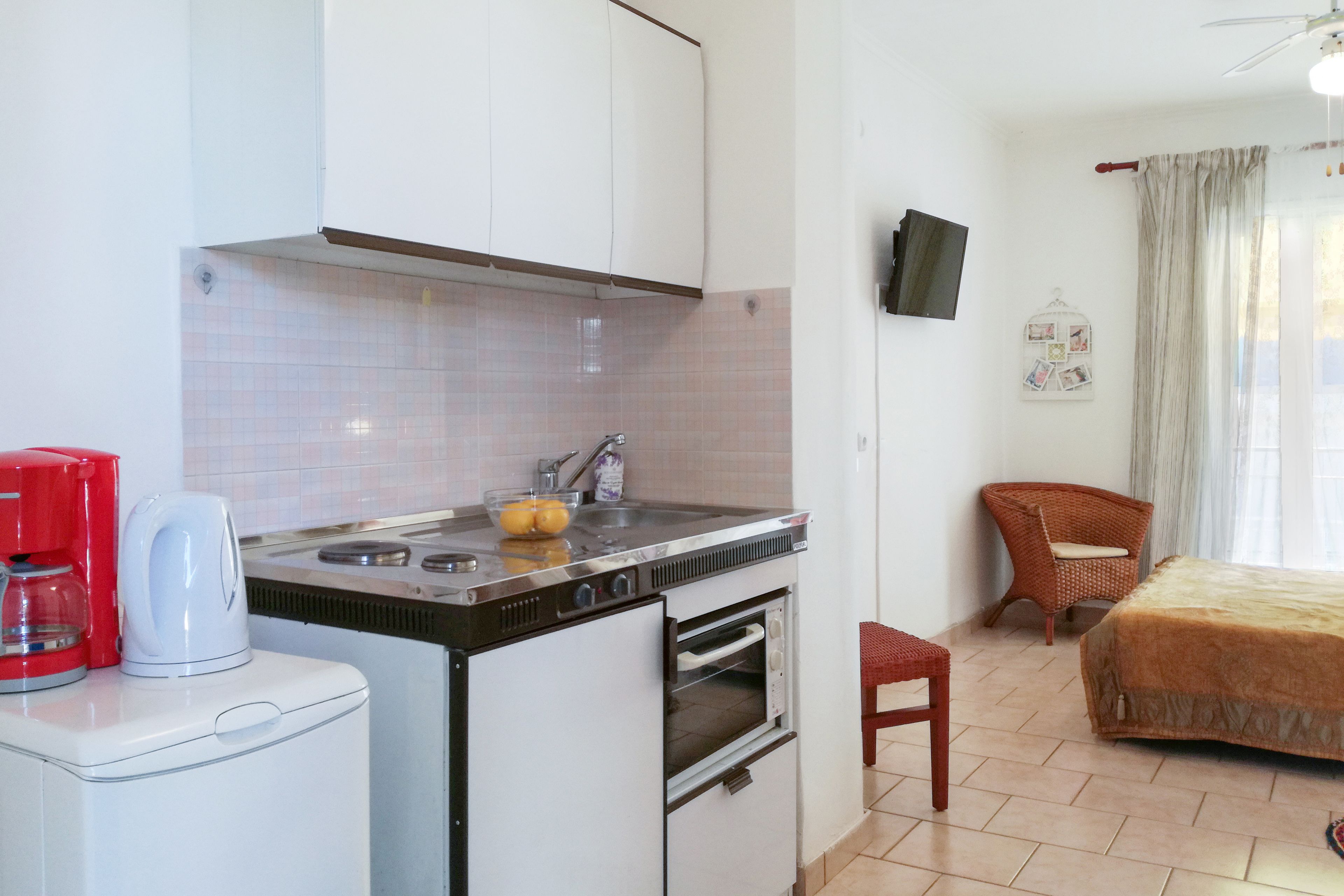 Cocina compacta con estufa, lavamanos y horno pequeño. Hay una cafetera roja y un tazón con limones en la encimera. Una silla de mimbre está cerca de una cama con manta color beige.