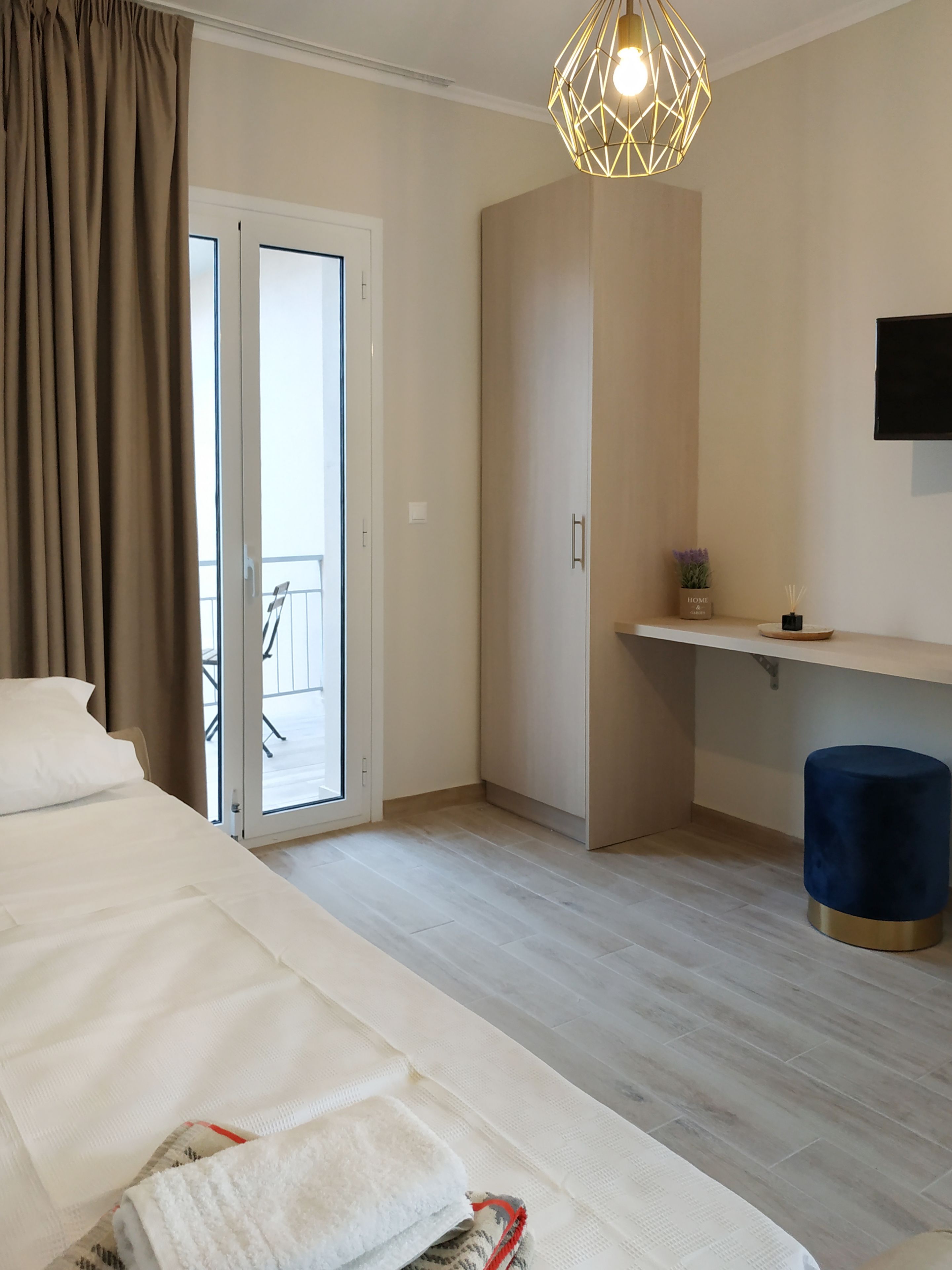 Une chambre d’hôtel avec un lit, un bureau, une chaise, un pouf bleu, une plante en pot et un téléviseur fixé au mur.