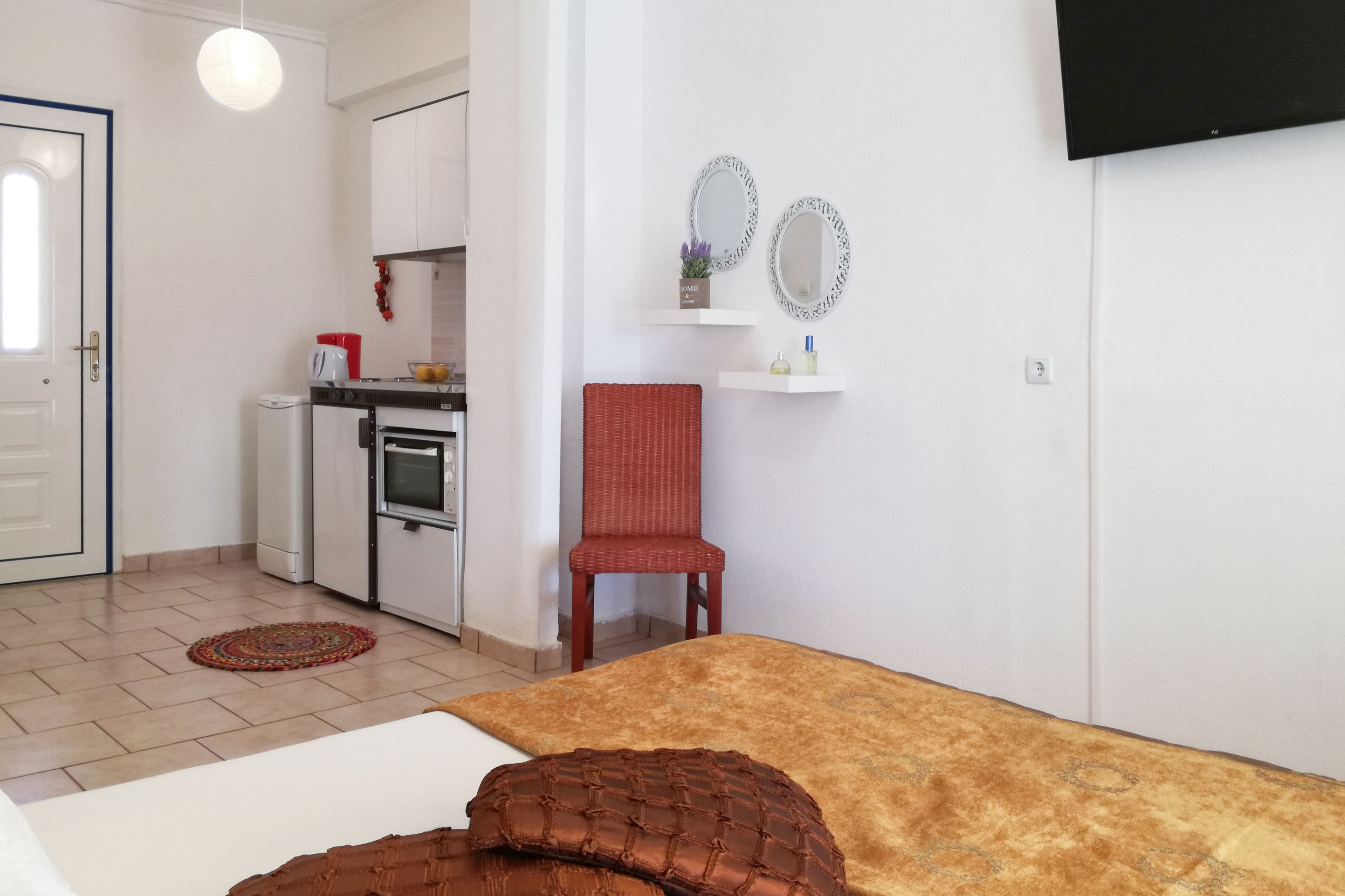 A compact room with a bed, a red chair, a kitchenette, and a TV mounted on the wall.