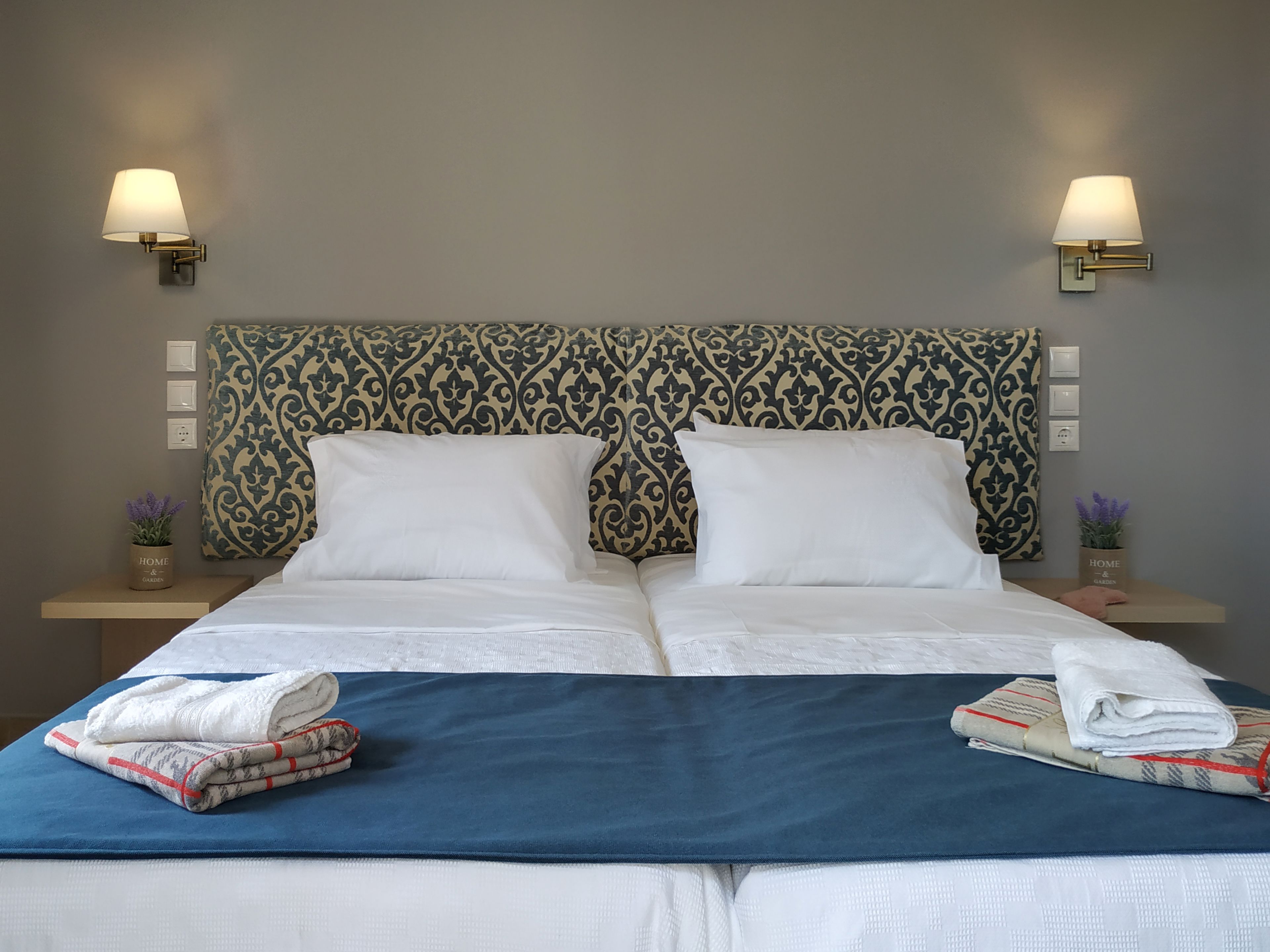 Un lit double bien fait, avec du linge de lit blanc, une couvre-lit bleue et deux tables de chevet ornées de lampes et de petites plantes en pot.