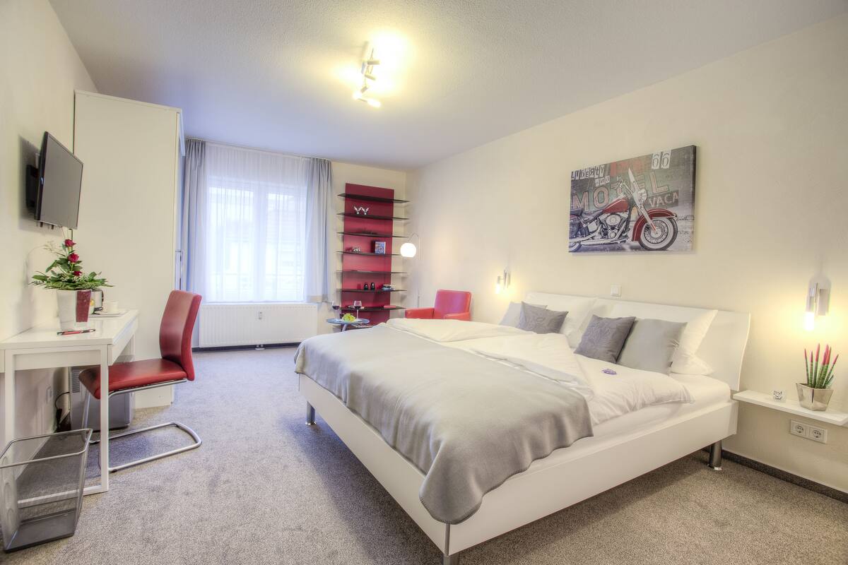 A modern hotel room with a large bed, a desk, a chair, a TV, and a motorcycle painting on the wall.