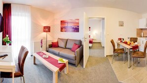 A modern living room with a sofa, a dining table with chairs, a kitchen area with a microwave, and a TV mounted on the wall. - Wehyer Aparthotel (Weyhe)