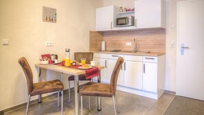 A compact kitchen with a microwave, a sink, and cabinets. There is a dining table with chairs and breakfast items like orange juice and a croissant. - Wehyer Aparthotel (Weyhe)
