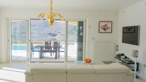 A modern living room with a white sofa, a dining table with chairs, a chandelier, and a view of a pool and mountains through large glass doors. - Villa Nika (Pag)