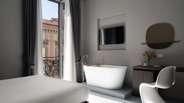 A modern hotel room with a large bed, a freestanding bathtub, a minimalist vanity, and a large window offering a view of the city. - The Grey (Naples)