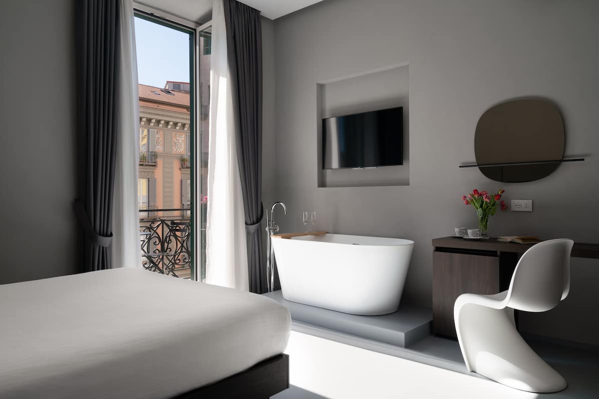 A modern hotel room with a large bed, a freestanding bathtub, a minimalist vanity, and a large window offering a view of the city.