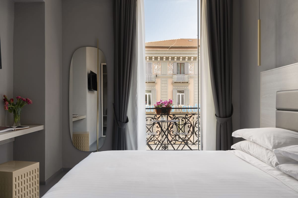 A hotel room with a large bed, a bedside table, a mirror, and a view of a building through a window with curtains.
