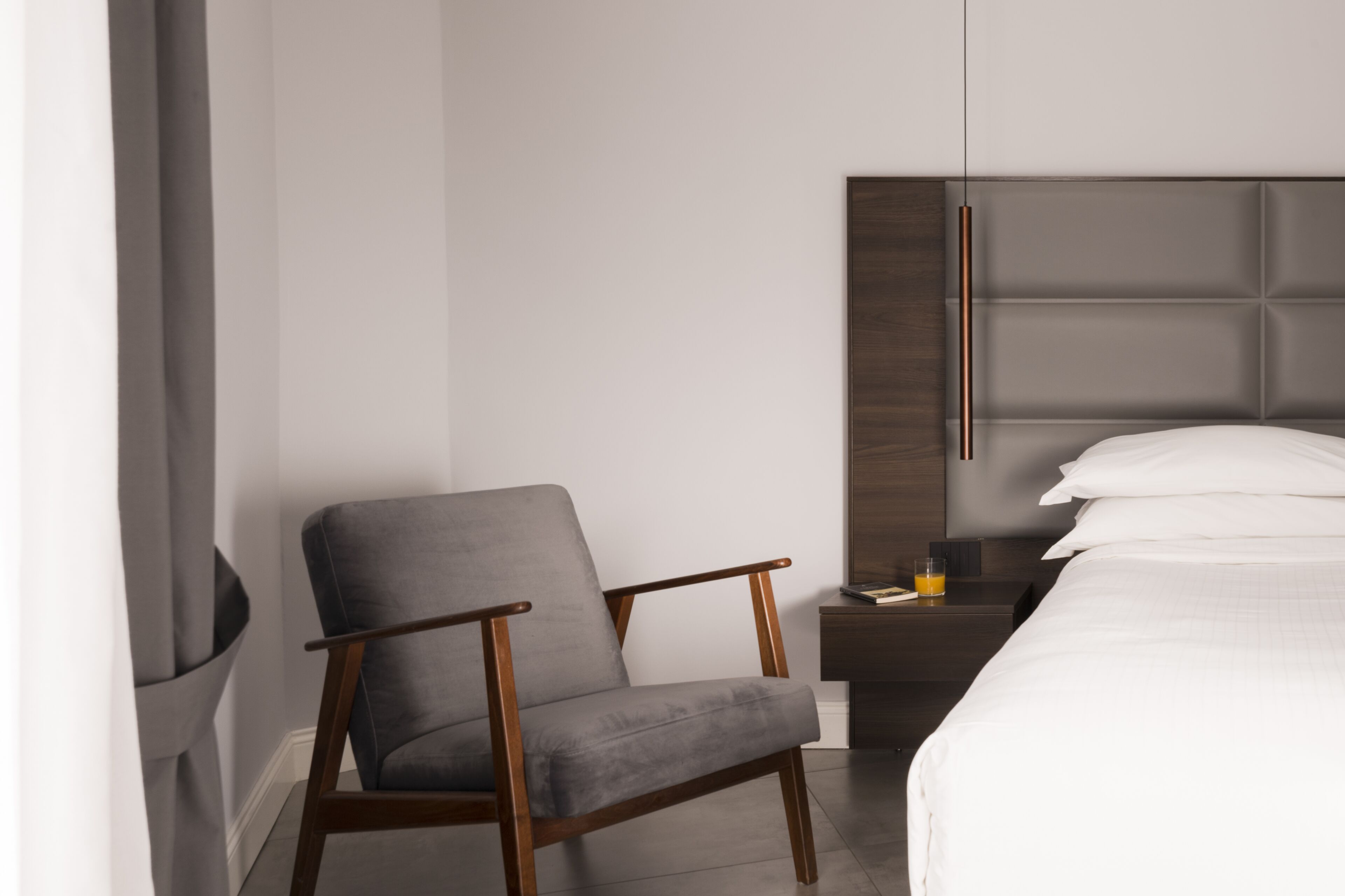 A modern hotel room with a grey upholstered armchair, a wooden nightstand with a book and a glass of orange juice, and a bed with white linens.
