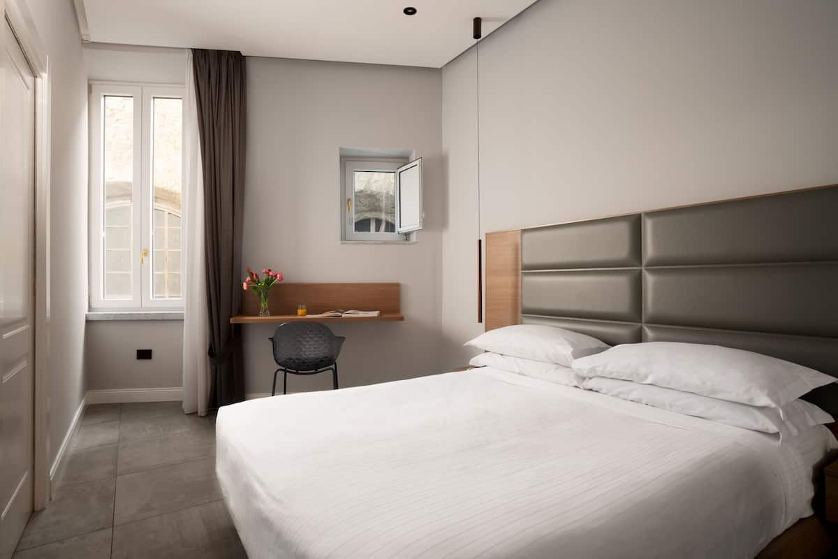 A hotel room with a bed, a desk, a chair, a window with curtains, and a small mirror on the wall.