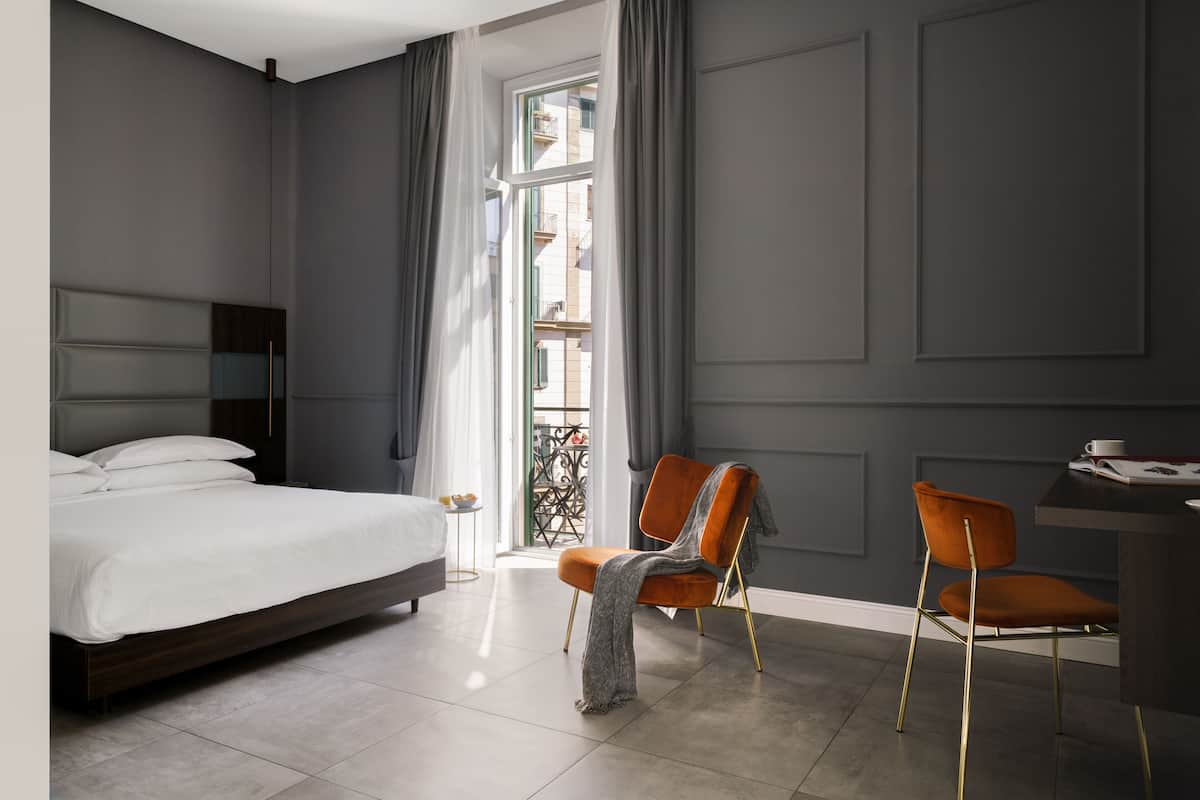 A modern hotel room with a large bed, two chairs, a desk, and a view of the outside through a window.