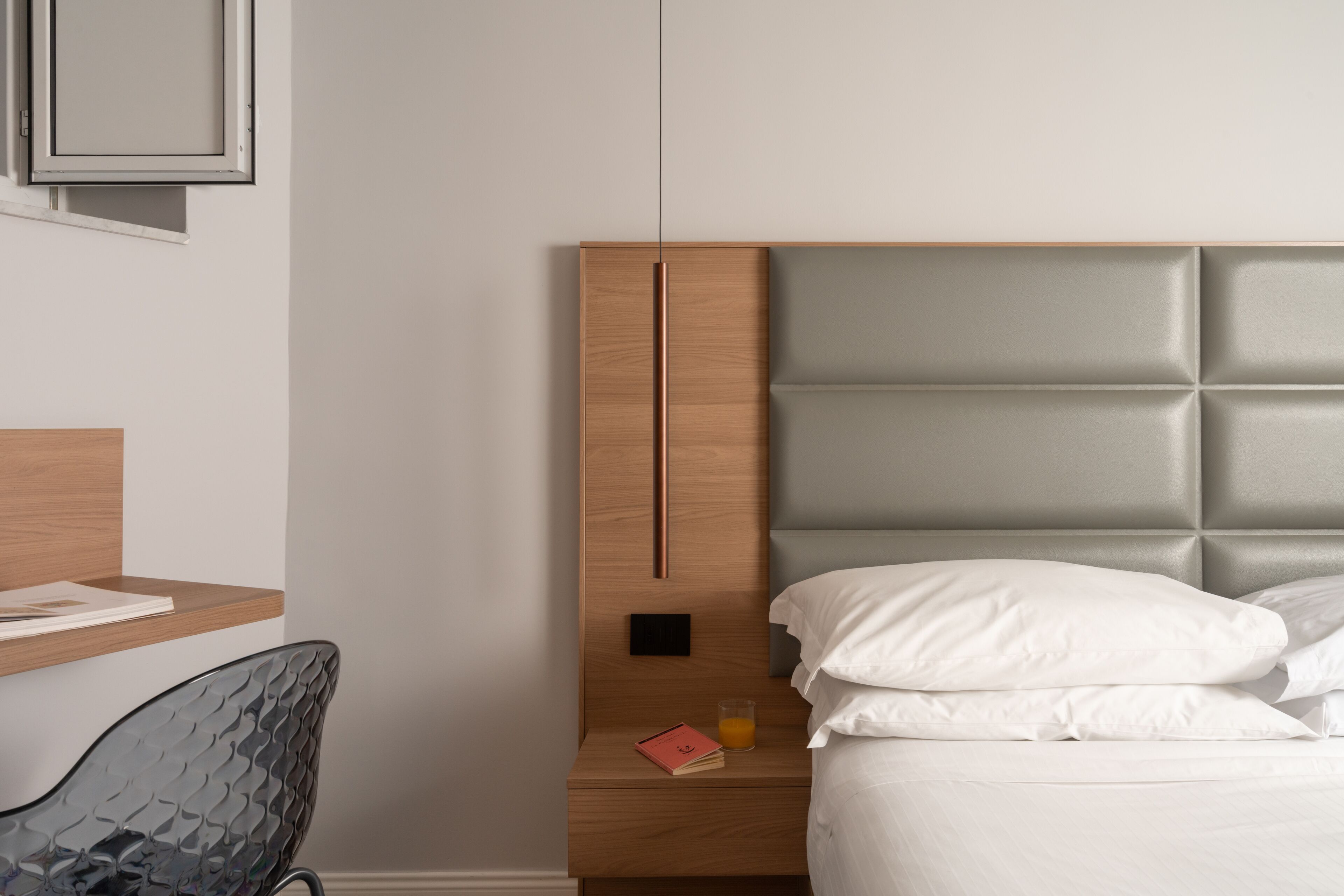 A modern bedroom with a wooden headboard, a glass chair, a bedside table with a book and a glass of orange juice, and a bed with white bedding.