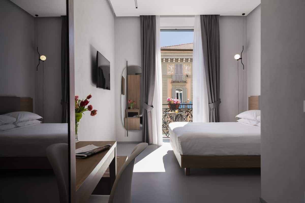 A modern hotel room with two beds, a TV, a desk, and a balcony with a view of the city.