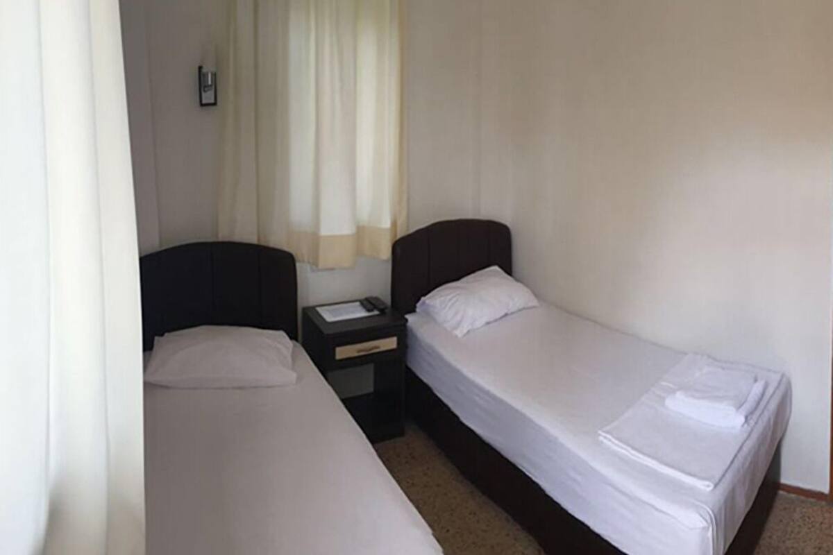 Une petite chambre propre, avec deux lits simples, une table de chevet et une fenêtre avec des rideaux.