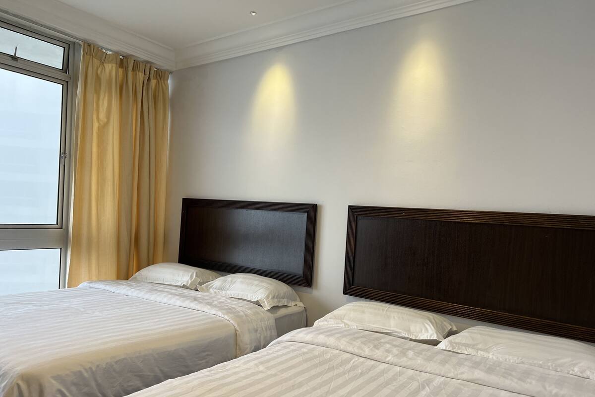 Ein Hotelzimmer mit zwei Betten, einem großen Fenster mit Vorhängen und einer schlichten Wand mit einer Lampe.