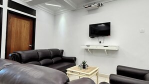 Una sala de estar moderna con un televisor de pantalla plana montado en una pared blanca, sofás de cuero negro, una mesa de centro de madera e iluminación empotrada con diseños geométricos en el techo.