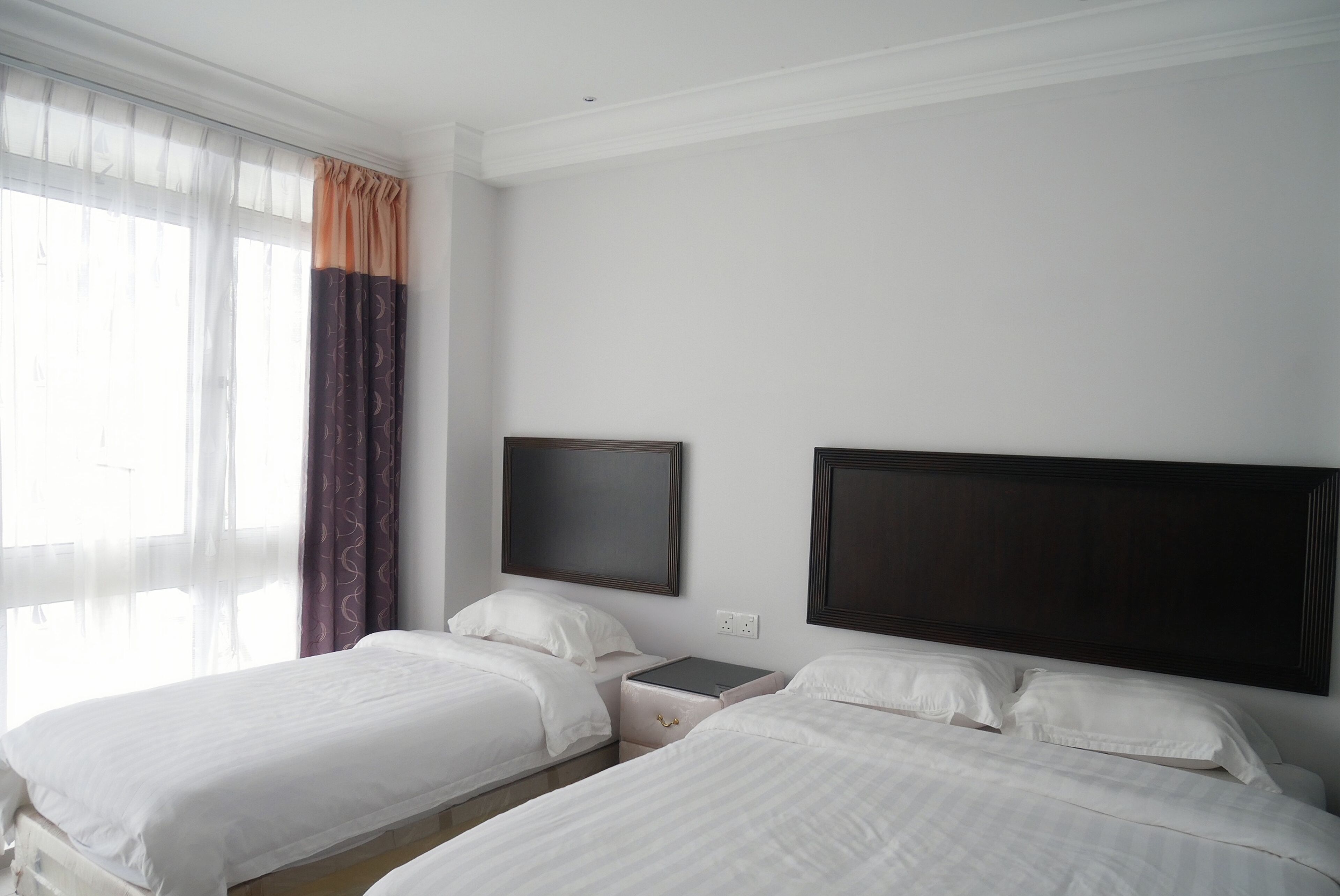 Habitación de hotel con dos camas, una mesita de noche, un televisor de pantalla plana y una ventana con cortinas translúcidas.
